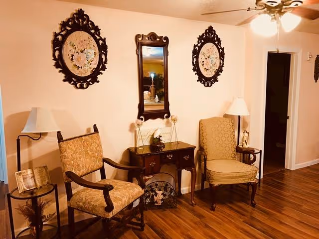 A sitting area with two upholstered armchairs flanking a small wooden table topped with decorations, a wall mirror and framed floral plaques, and hardwood floors.
