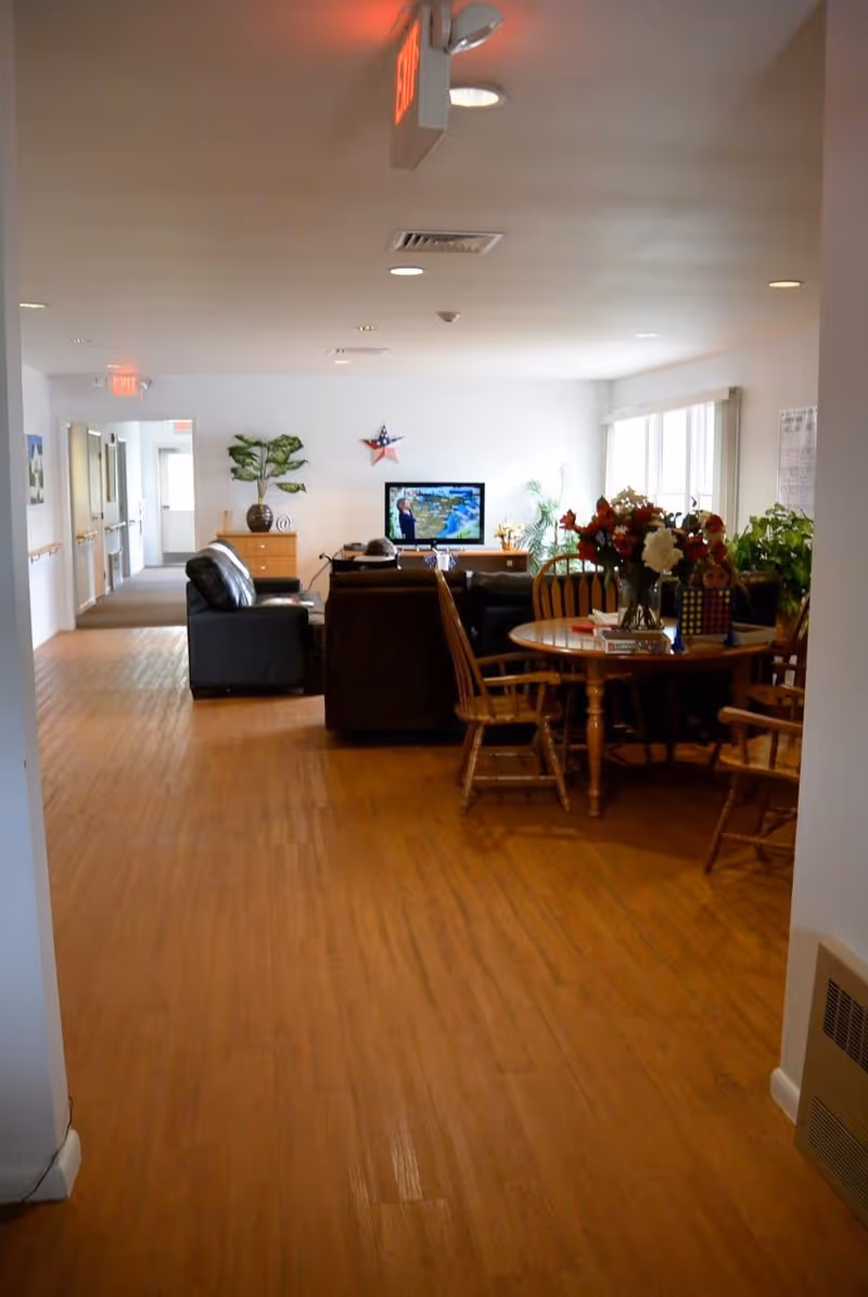 Interior view of a senior living facility common area with wooden flooring, a round wooden table with chairs and a vase of flowers, several armchairs facing a television mounted on a white wall, and plants placed around the room. There are exit signs on the ceiling and a hallway leading to other rooms.