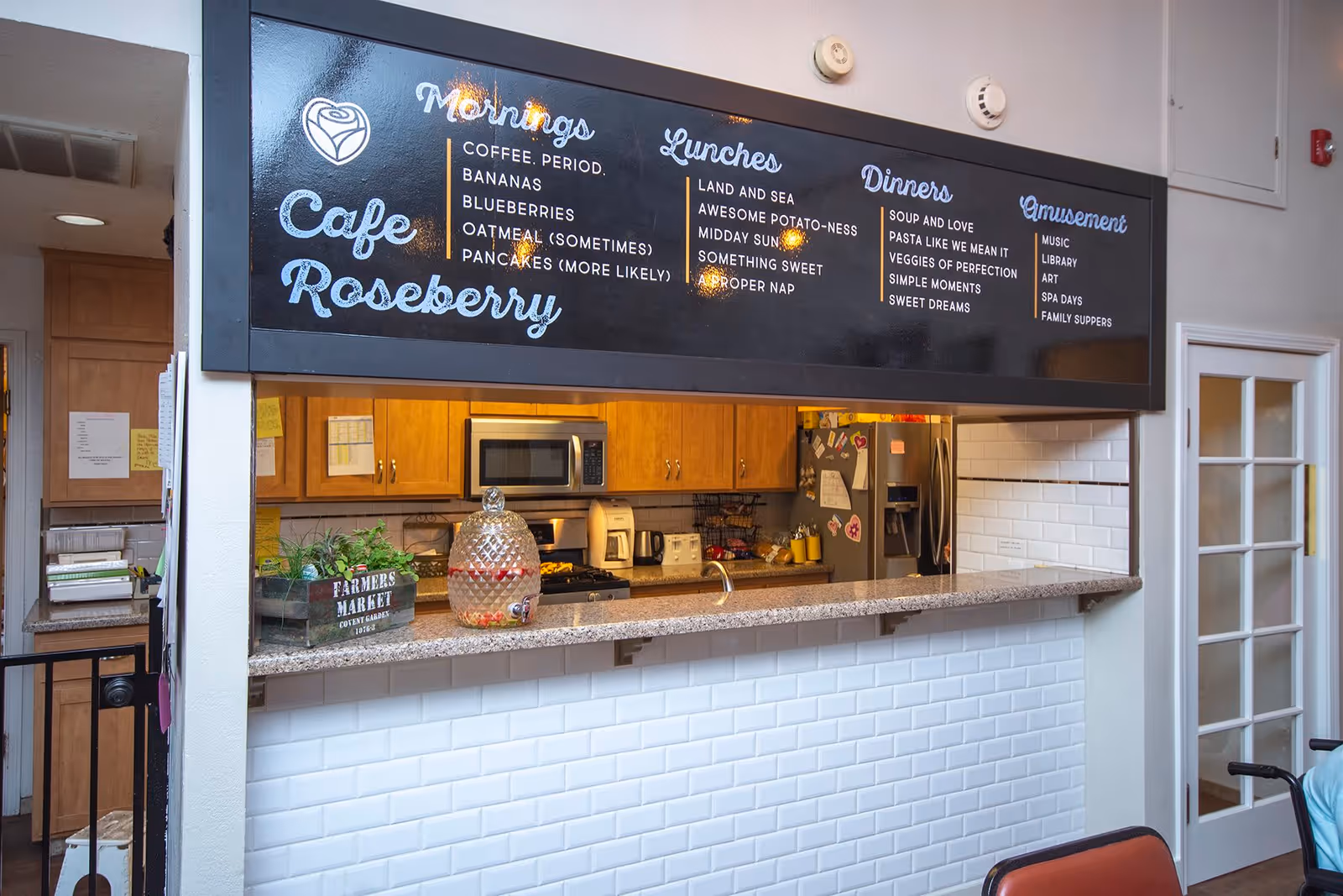 Interior view of Cafe Roseberry with a serving counter and a large black menu board above it listing options for mornings, lunches, dinners, and amusement activities. The kitchen area behind the counter has wooden cabinets, a microwave, a refrigerator with magnets, and various kitchen appliances. A glass door is visible to the right, and a small plant and a beverage dispenser are on the counter.