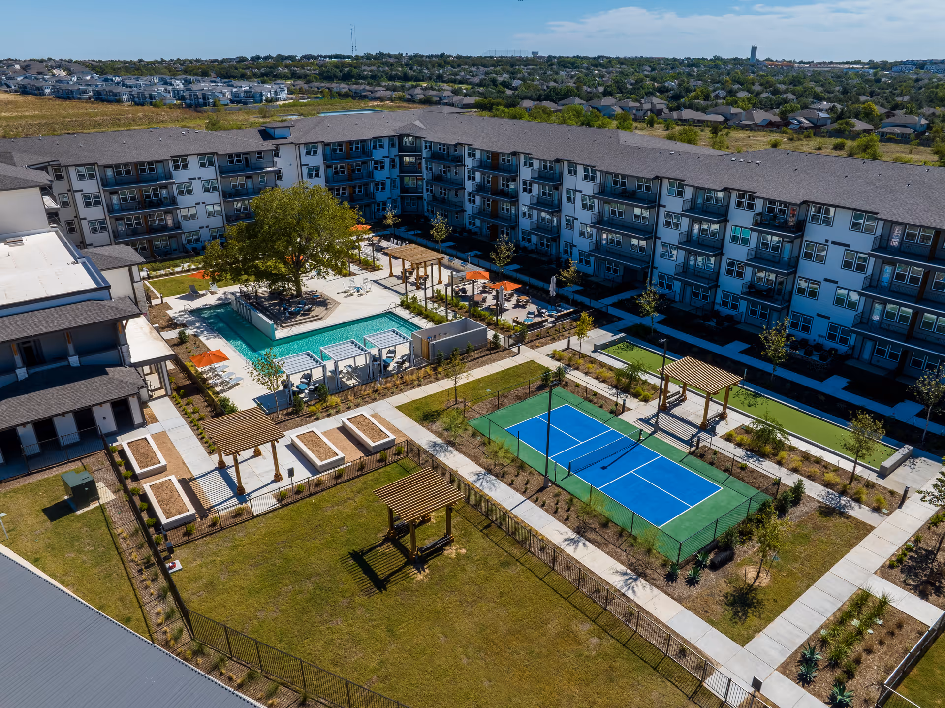 Aerial view of Amberlin Georgetown senior living facility showing a large multi-story residential building surrounding an outdoor courtyard with a swimming pool, lounge chairs, pergolas, a blue pickleball court, and landscaped green areas with pathways.