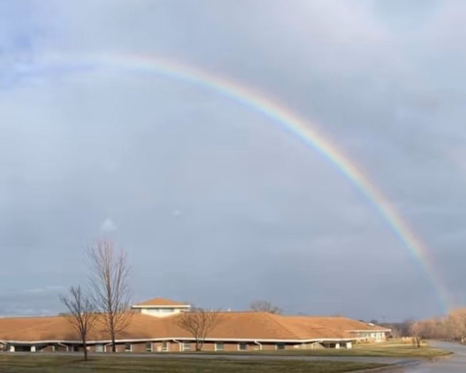 A wide exterior view of a single-story building with a brown roof under a cloudy sky, featuring a vibrant rainbow arching above it. There are a few leafless trees in front of the building and a paved road to the right.