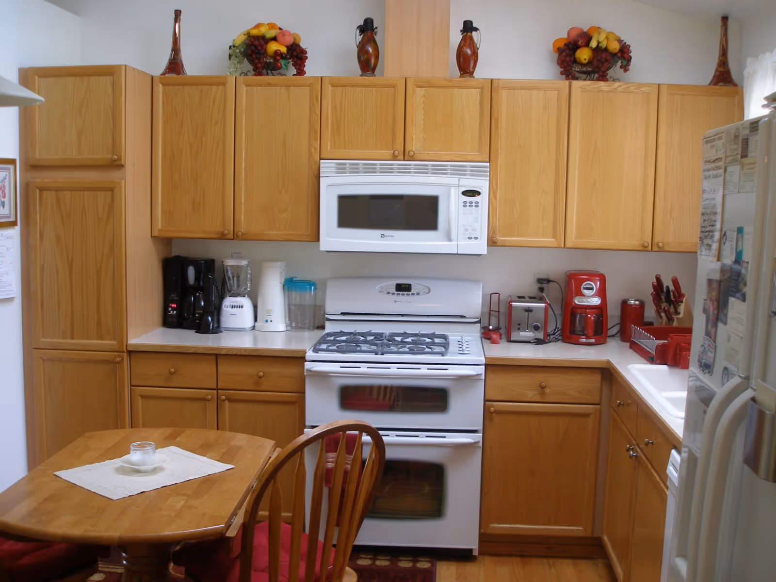 A kitchen with wooden cabinets, a white gas stove and oven, a white microwave above the stove, and various small appliances including a coffee maker, blender, toaster, and red coffee machine on the countertops. There is a wooden dining table with a white placemat and a small candle, surrounded by wooden chairs with red cushions. Decorative fruit baskets and vases are placed on top of the cabinets.