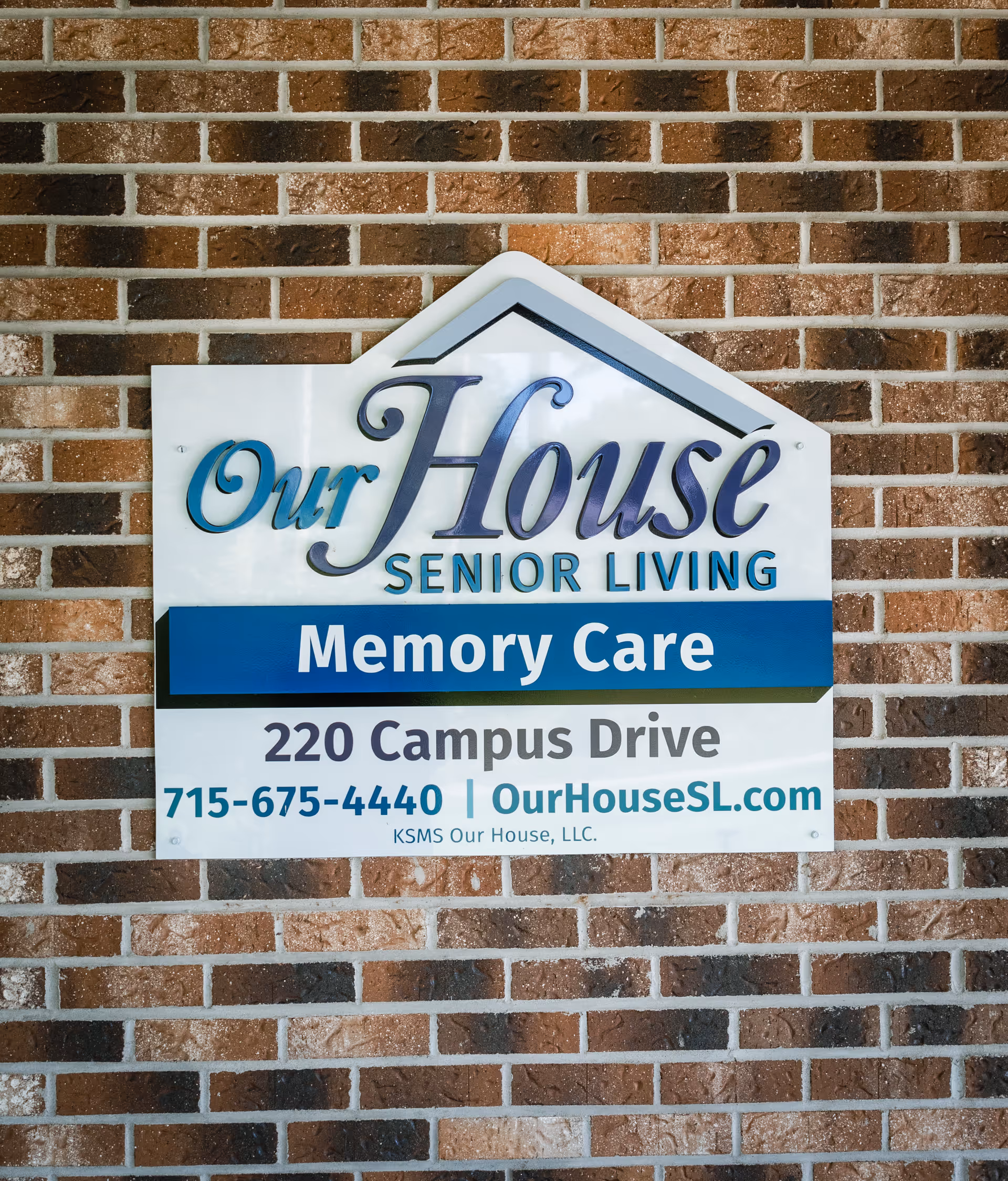 A sign mounted on a brick wall that reads 'Our House Senior Living Memory Care' with the address 220 Campus Drive, phone number 715-675-4440, and website OurHouseSL.com.