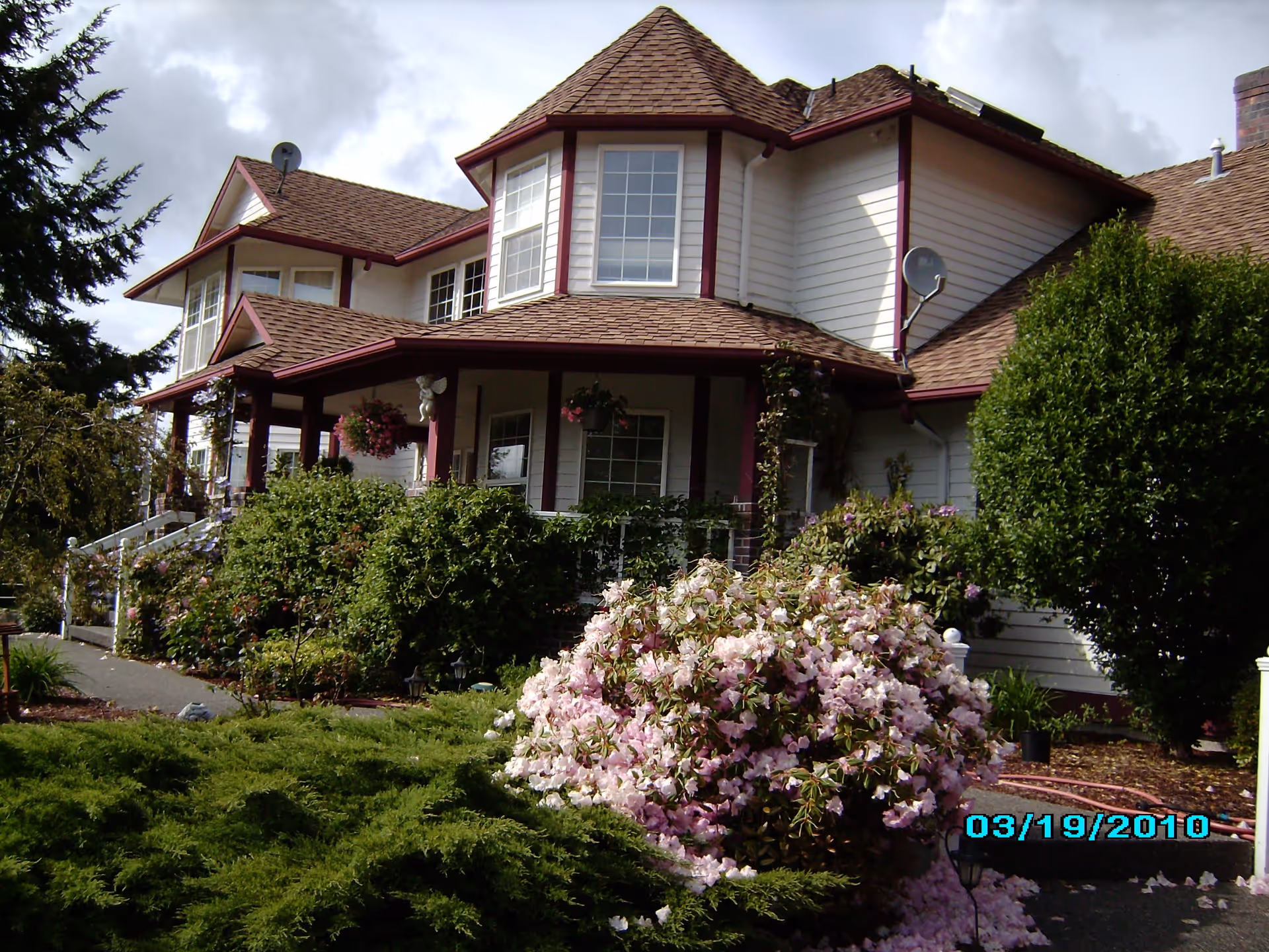 Exterior view of a large two-story house with white siding and brown roof shingles. The house has multiple windows and a covered porch with hanging flower baskets. The front yard is landscaped with green bushes and blooming pink flowers. The sky is cloudy.