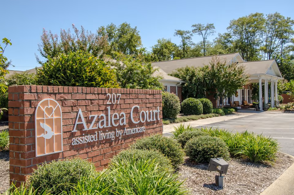Brick sign reading "Azalea Court assisted living by Americare" in front of a landscaped entrance and building with white columns.