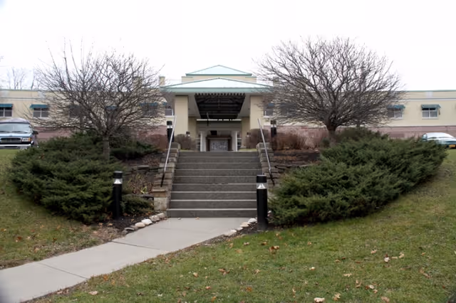 Front entrance of a low-rise building with a central staircase, handrails, shrubs, and leafless trees on a grassy slope.