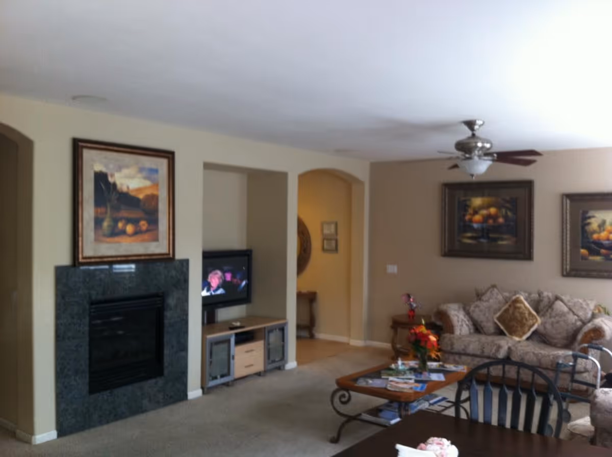 A cozy living room with a beige carpet and walls. There is a fireplace with a dark green marble surround and a framed painting above it. Next to the fireplace is a small TV on a wooden stand. The room has a ceiling fan with lights, a patterned sofa with decorative pillows, a wooden coffee table with magazines and a flower vase, and framed artwork on the walls. A dining table with chairs is partially visible in the foreground.