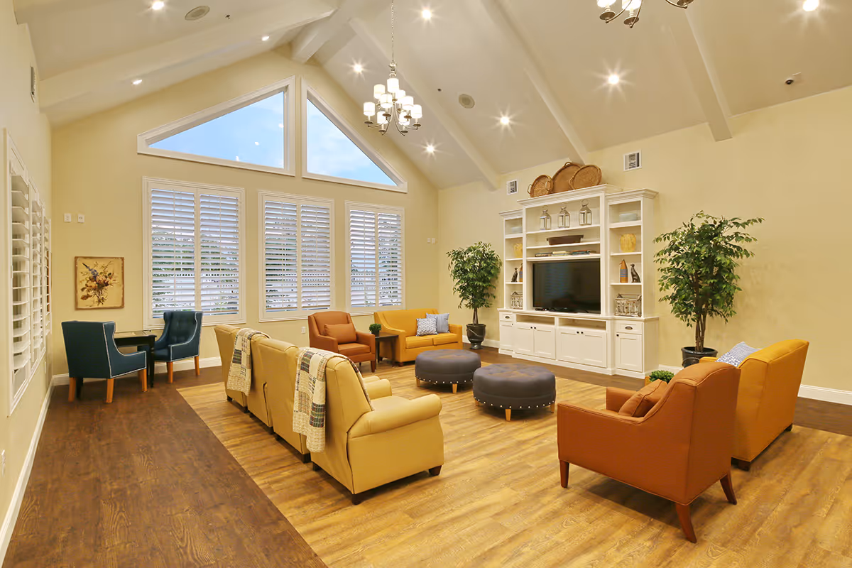 A spacious and well-lit living room with high vaulted ceilings and large triangular windows. The room features a seating arrangement with yellow and orange armchairs and sofas around two round ottomans. There is a white entertainment center with a TV and decorative items, flanked by two large potted plants. The floor is a combination of wood and laminate, and there are two blue chairs with a small table in the corner near the windows with white shutters.