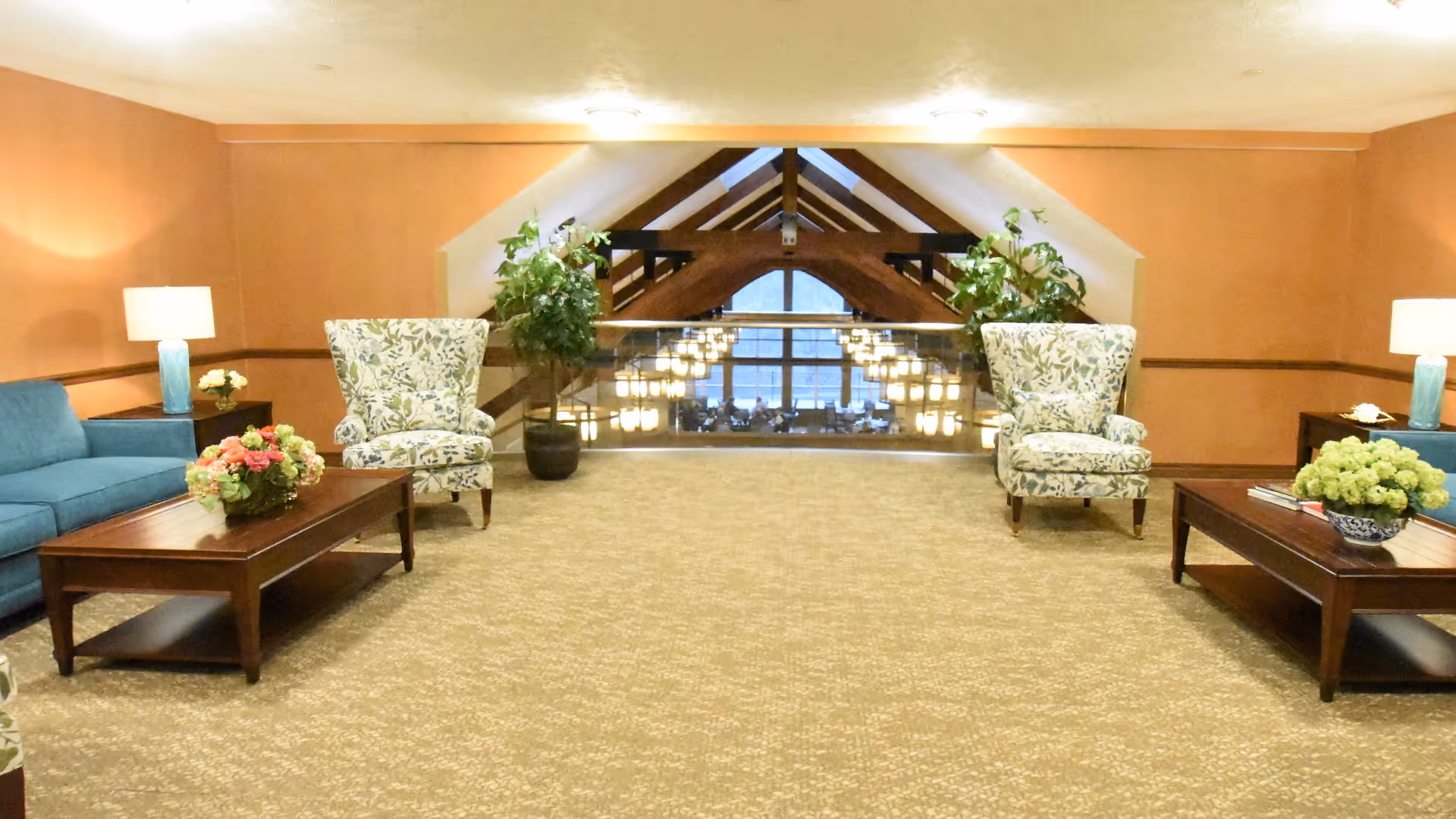 Lounge area with patterned armchairs, sofas, coffee tables, lamps and plants overlooking an atrium with hanging lights.