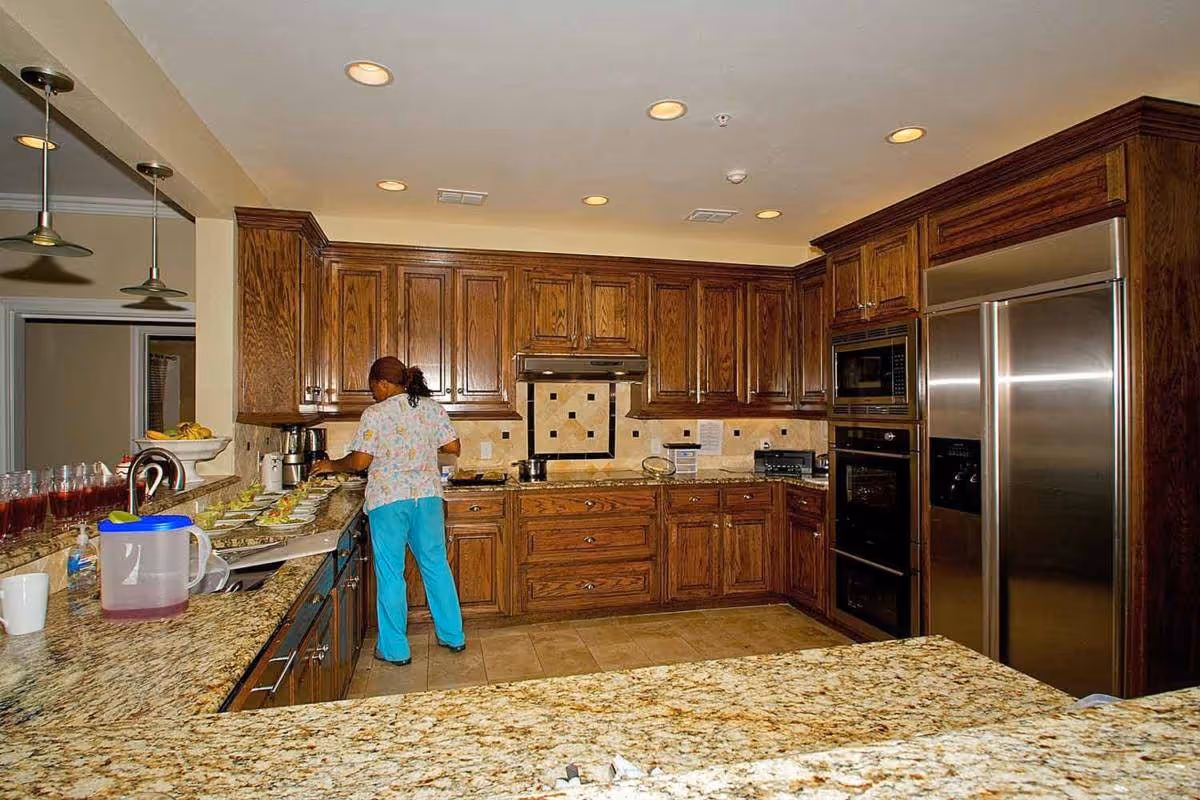 A kitchen area with wooden cabinets, granite countertops, and stainless steel appliances. A person wearing blue pants and a patterned shirt is preparing food on the counter. Several glasses filled with a red beverage and a pitcher are also visible on the counter.