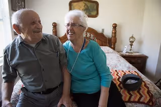An elderly man and woman sitting together on the edge of a bed in a bedroom, smiling and looking at each other. The room has a wooden bed frame, a bedside table with a lamp, and a colorful blanket folded at the foot of the bed.