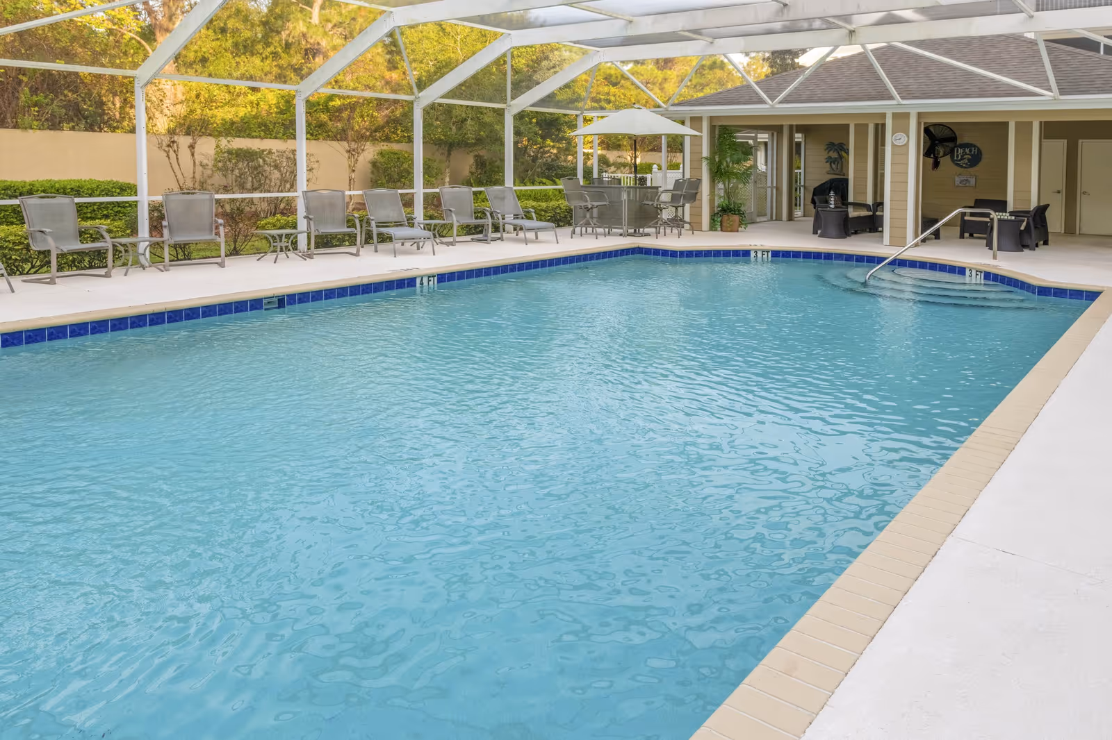 Indoor swimming pool with clear blue water surrounded by a white deck. Several lounge chairs and tables with umbrellas are arranged along the poolside. The area is enclosed with a screened roof and walls, with greenery visible outside the enclosure.