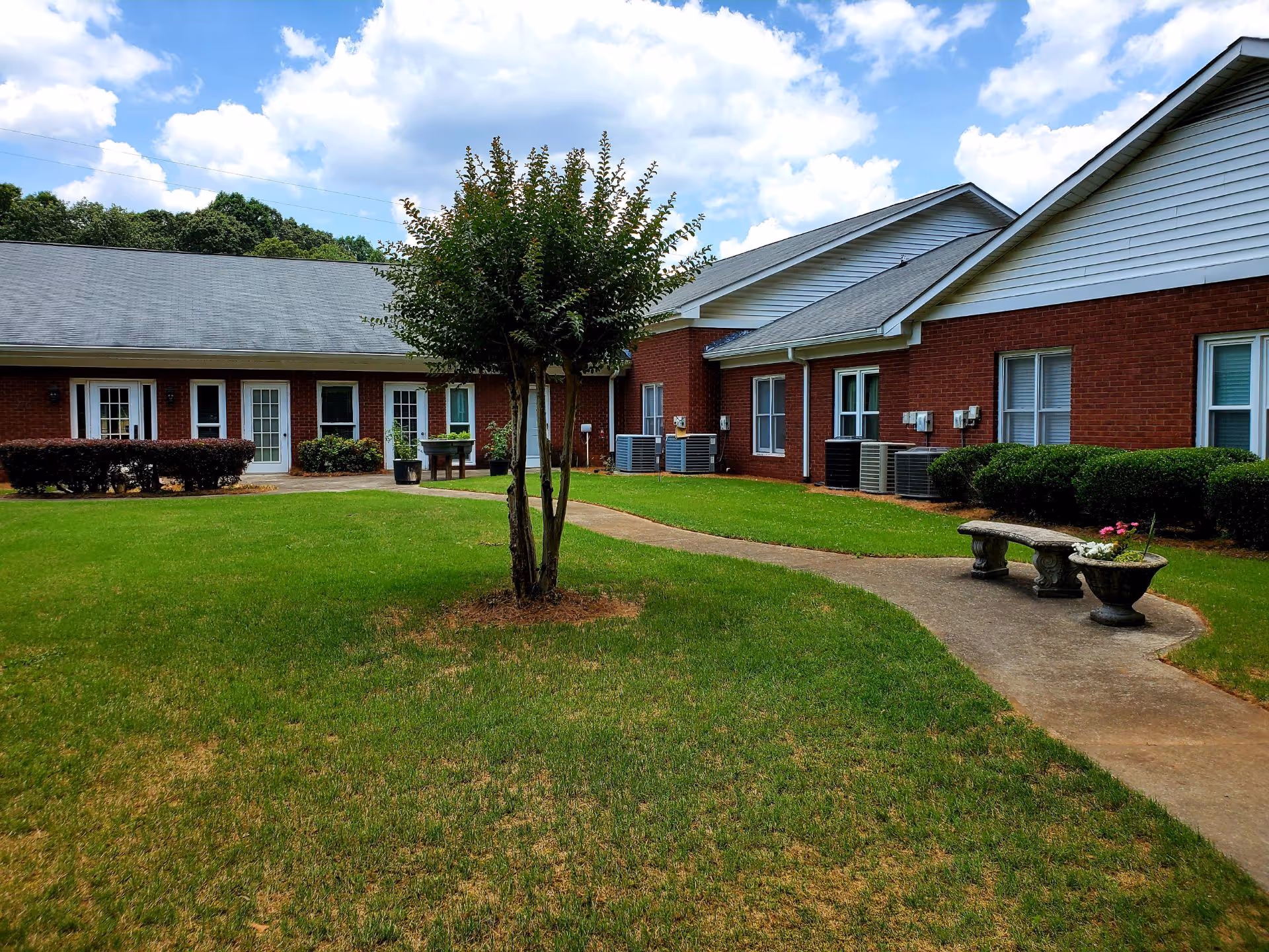 Outdoor courtyard area of a senior living facility with a green lawn, a small tree in the center, a concrete pathway, a stone bench, and flower pots. The building surrounding the courtyard has red brick walls, white doors, and windows with white trim under a gray shingled roof. The sky is partly cloudy.