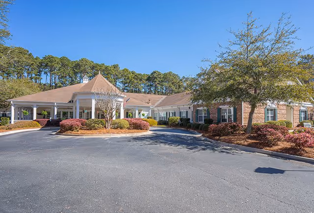 Exterior view of a single-story senior living facility building with a covered entrance, surrounded by landscaped bushes and trees under a clear blue sky.