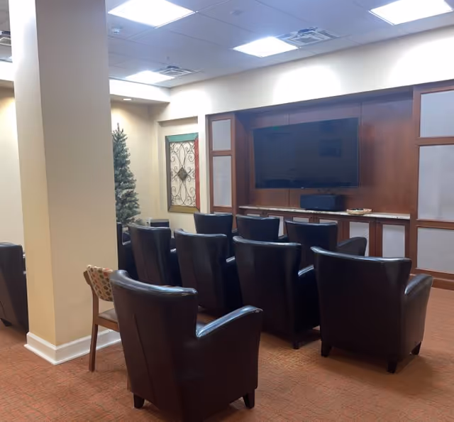 Leather armchairs arranged in rows facing a wall-mounted television in a carpeted common room with wood cabinetry and recessed ceiling lights.