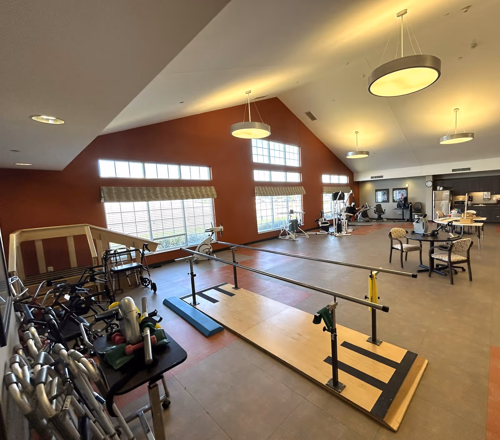 A spacious rehabilitation and exercise room with parallel bars for walking practice, various exercise equipment, walkers, and a table with chairs. The room has large windows with natural light, a high ceiling with hanging lights, and a kitchen area in the background.