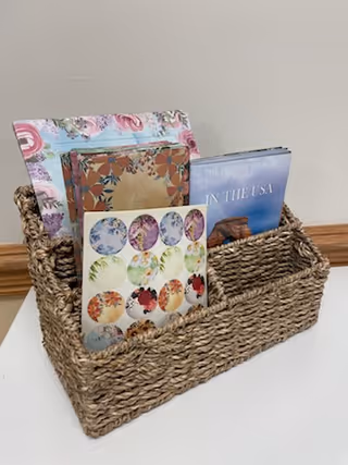 Woven rectangular basket on a white table against a beige wall, holding brochures, booklets, and greeting cards.