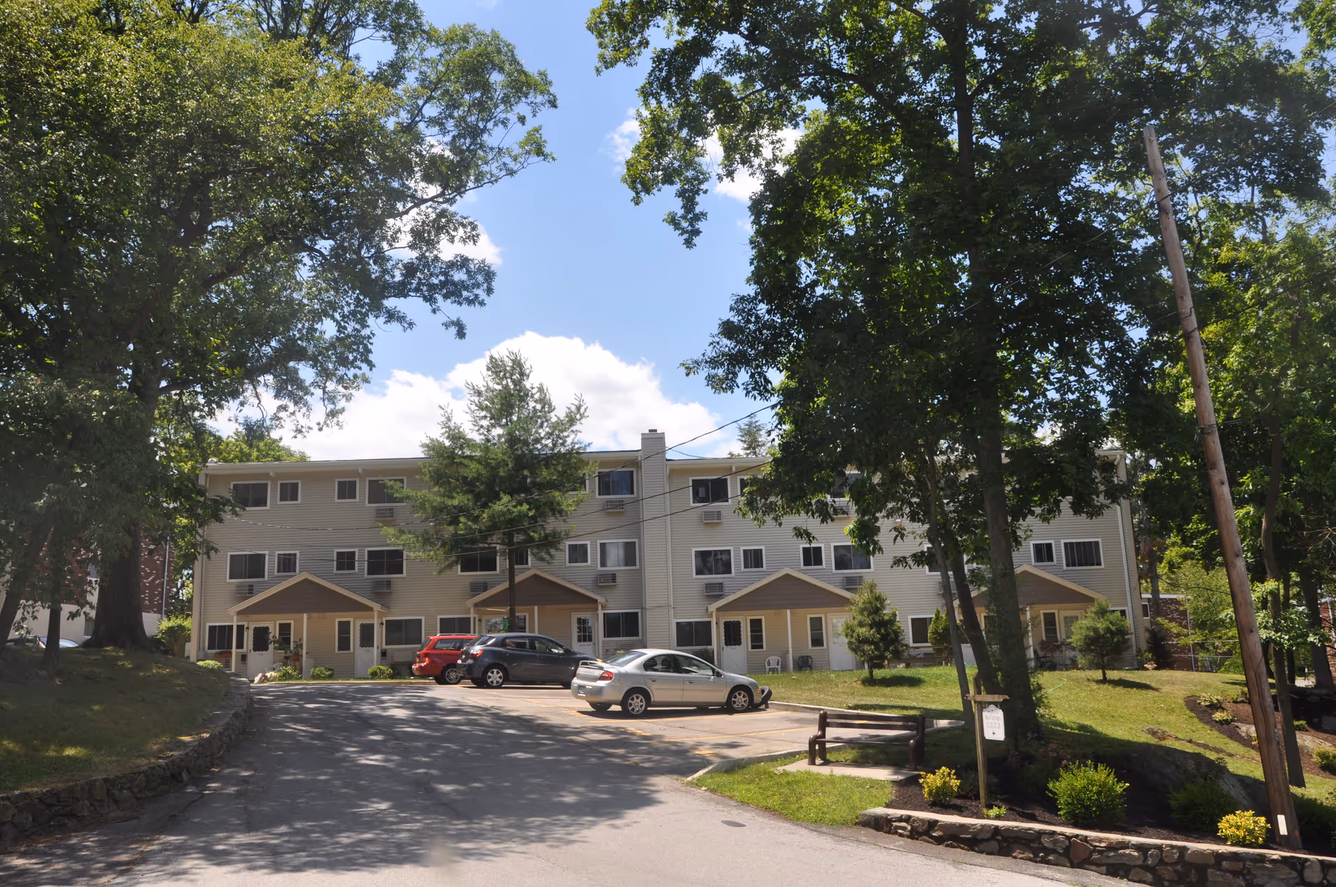 Three-story beige apartment building with parked cars, a driveway, and large trees in front.