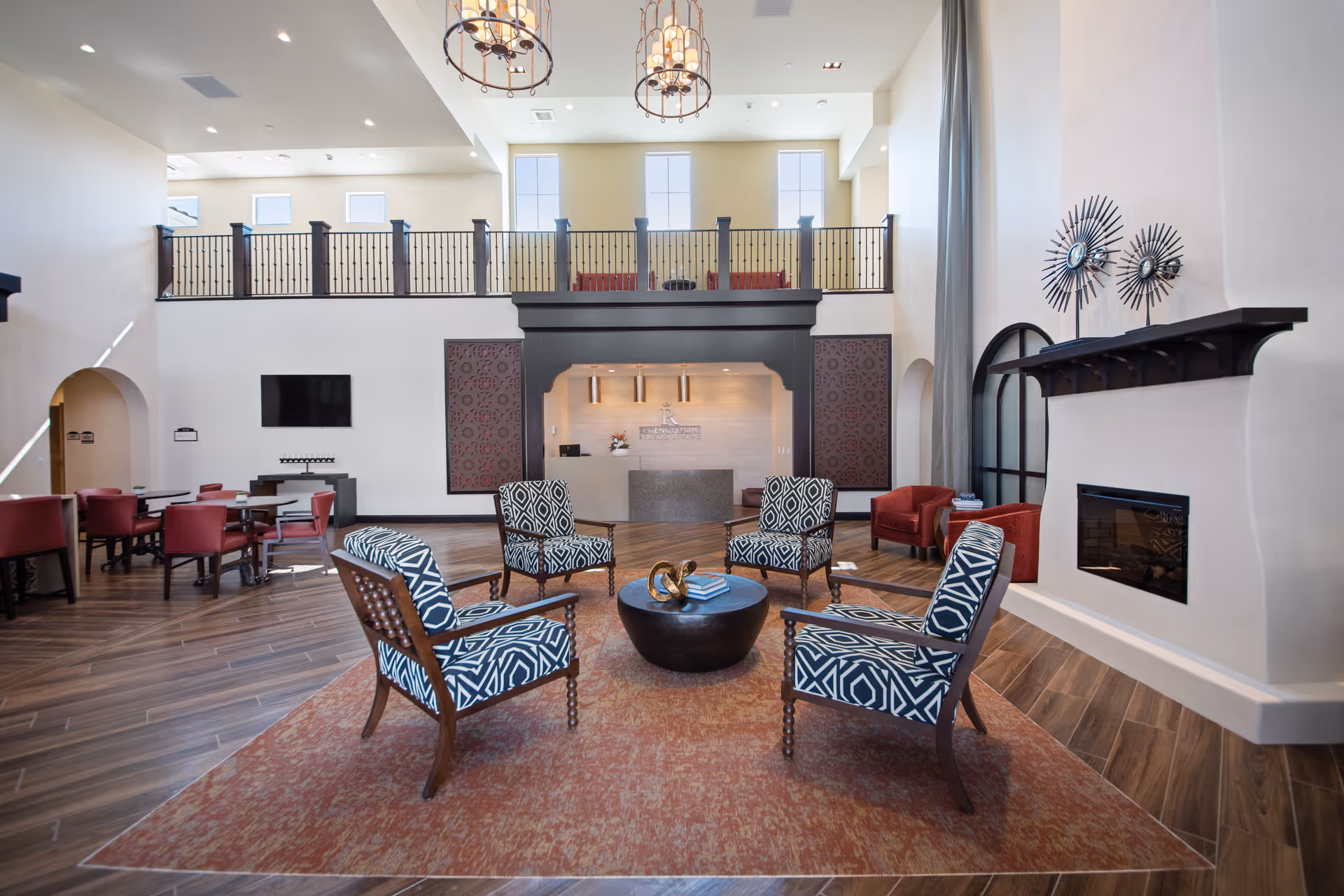 Spacious two-story lounge with patterned chairs around a central coffee table, fireplace, and additional seating under a balcony overlooking the lobby.