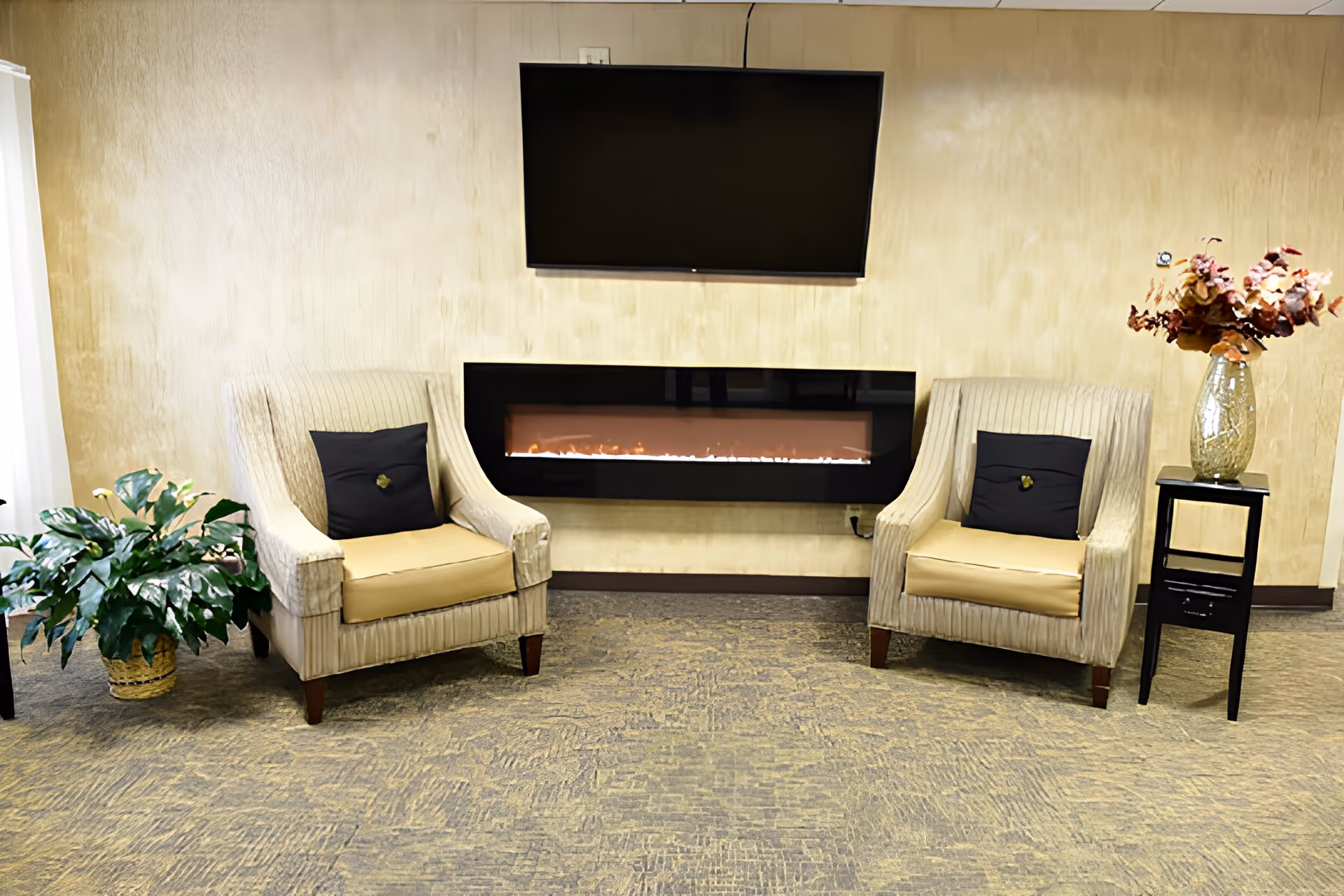 A cozy seating area with two beige armchairs, each with a black cushion, positioned on either side of a modern electric fireplace mounted on a beige wall. Above the fireplace is a flat-screen TV. To the left of the left chair is a green potted plant, and to the right of the right chair is a small black side table with a glass vase holding dried flowers.