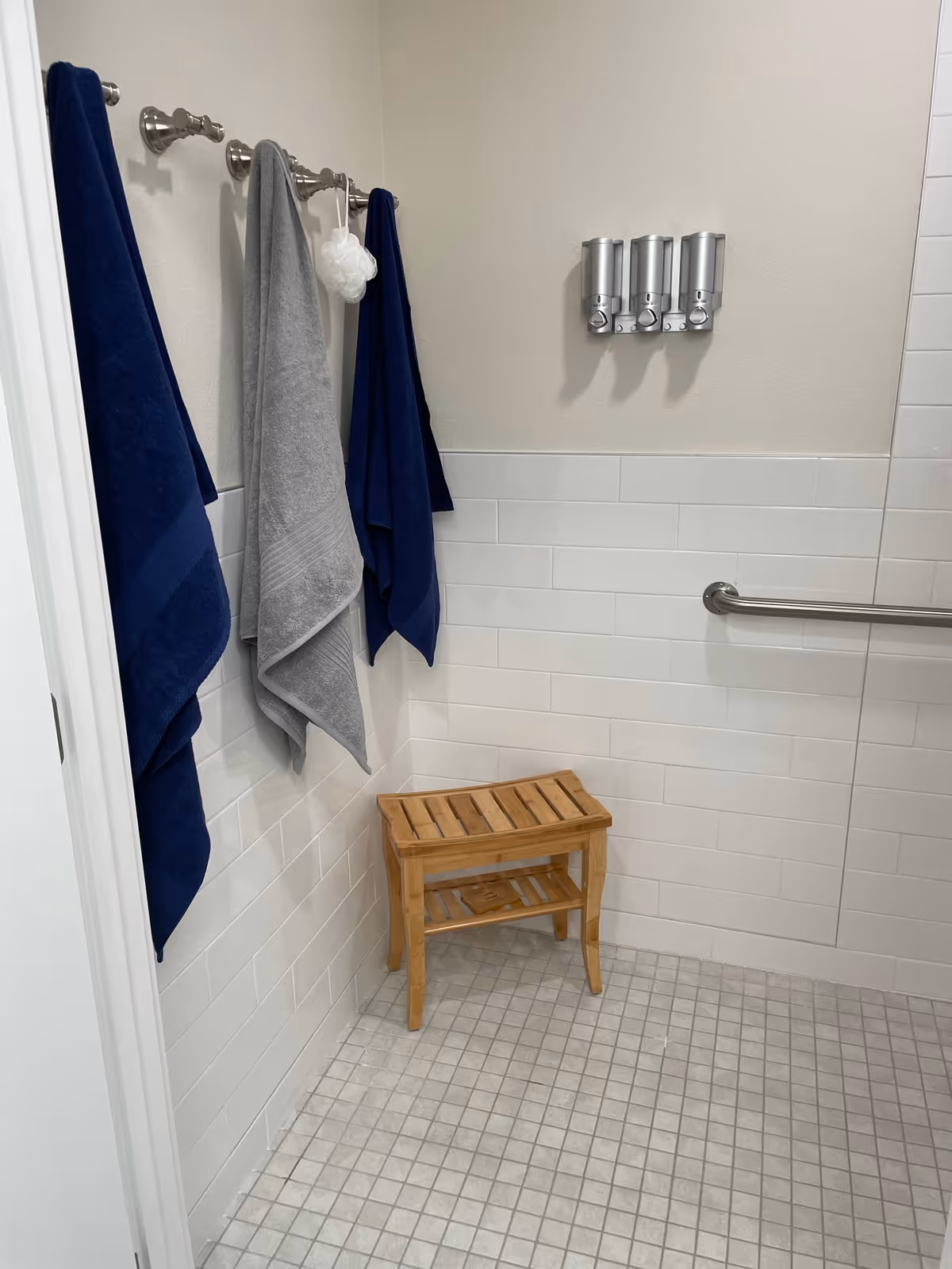 A bathroom corner with white tiled walls and floor, featuring three towels hanging on hooks (two navy blue and one gray), a wooden shower bench, a grab bar, and three wall-mounted soap or shampoo dispensers.