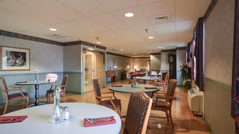 A dining room in a senior living facility with round tables set with napkins, cutlery, and small flower vases. The room has wooden chairs with cushions, framed artwork on the walls, a grandfather clock, and large windows with curtains letting in natural light.