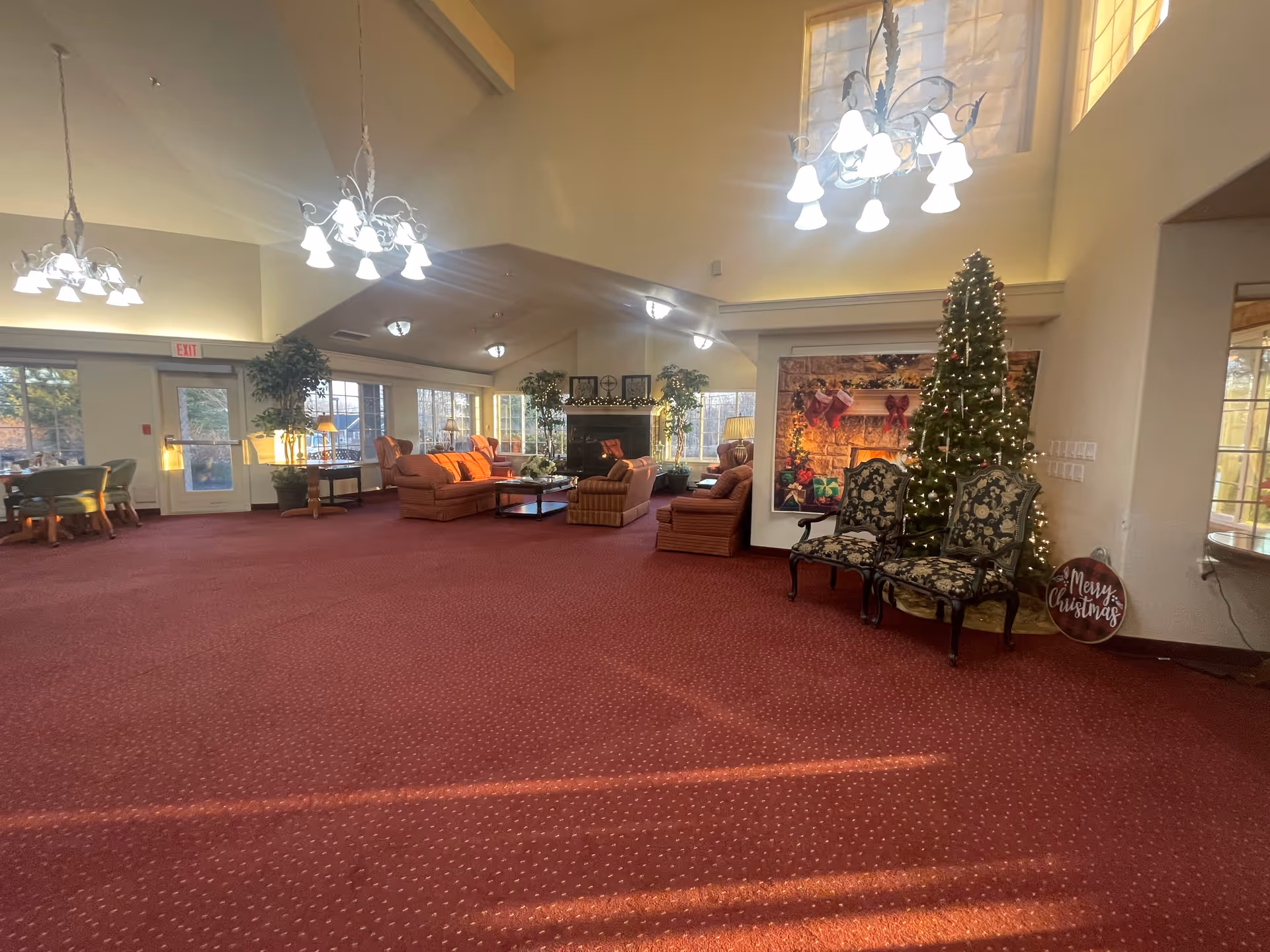 A spacious senior living community lounge area with red carpet flooring, multiple seating arrangements including sofas and armchairs, large windows letting in natural light, a decorated Christmas tree with lights, and a festive 'Merry Christmas' sign. The room features high ceilings with chandeliers and a cozy fireplace surrounded by plants and holiday decorations.