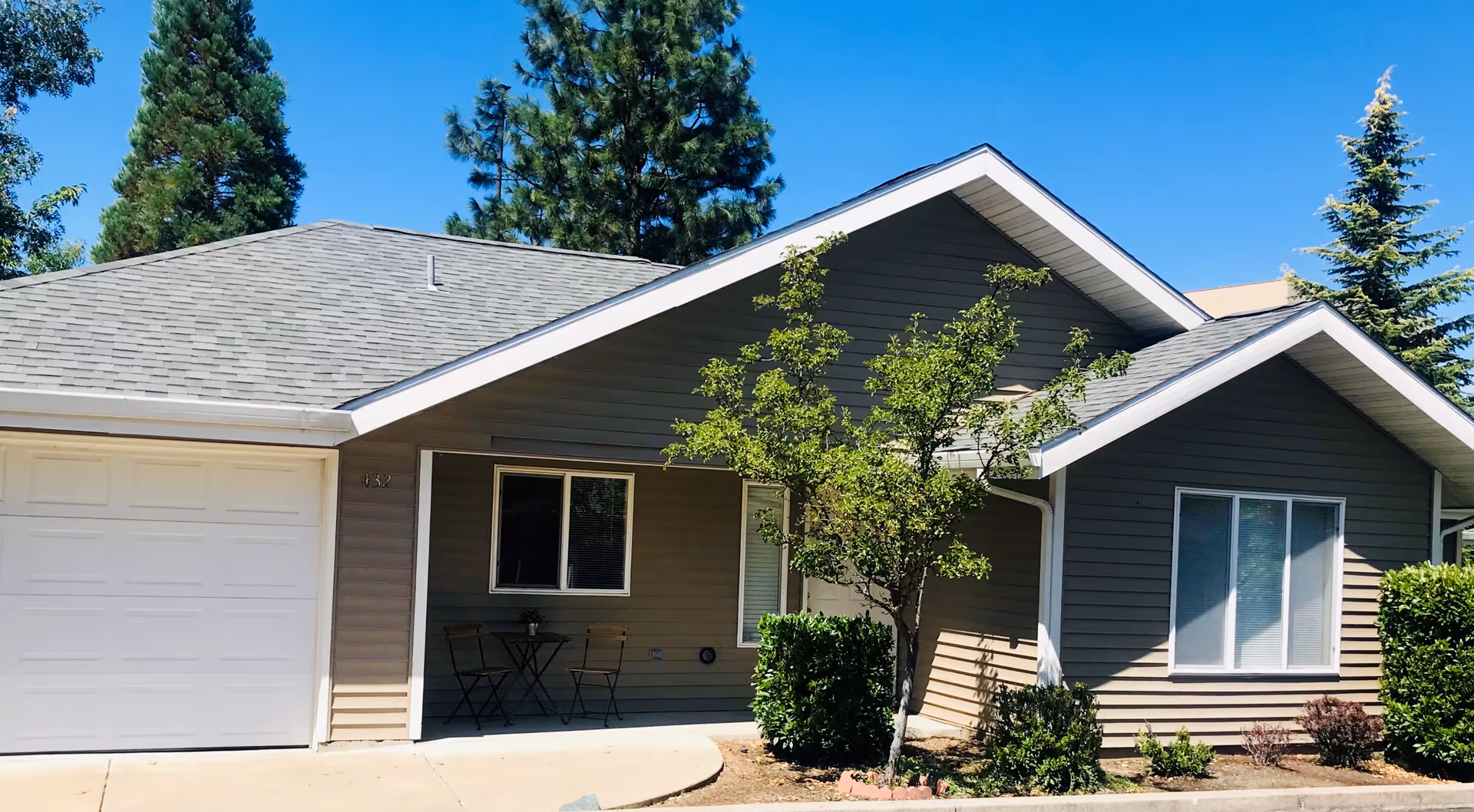Exterior view of a single-story residential building with gray siding and a white garage door. There is a small covered porch area with a table and two chairs, surrounded by small trees and bushes under a clear blue sky.