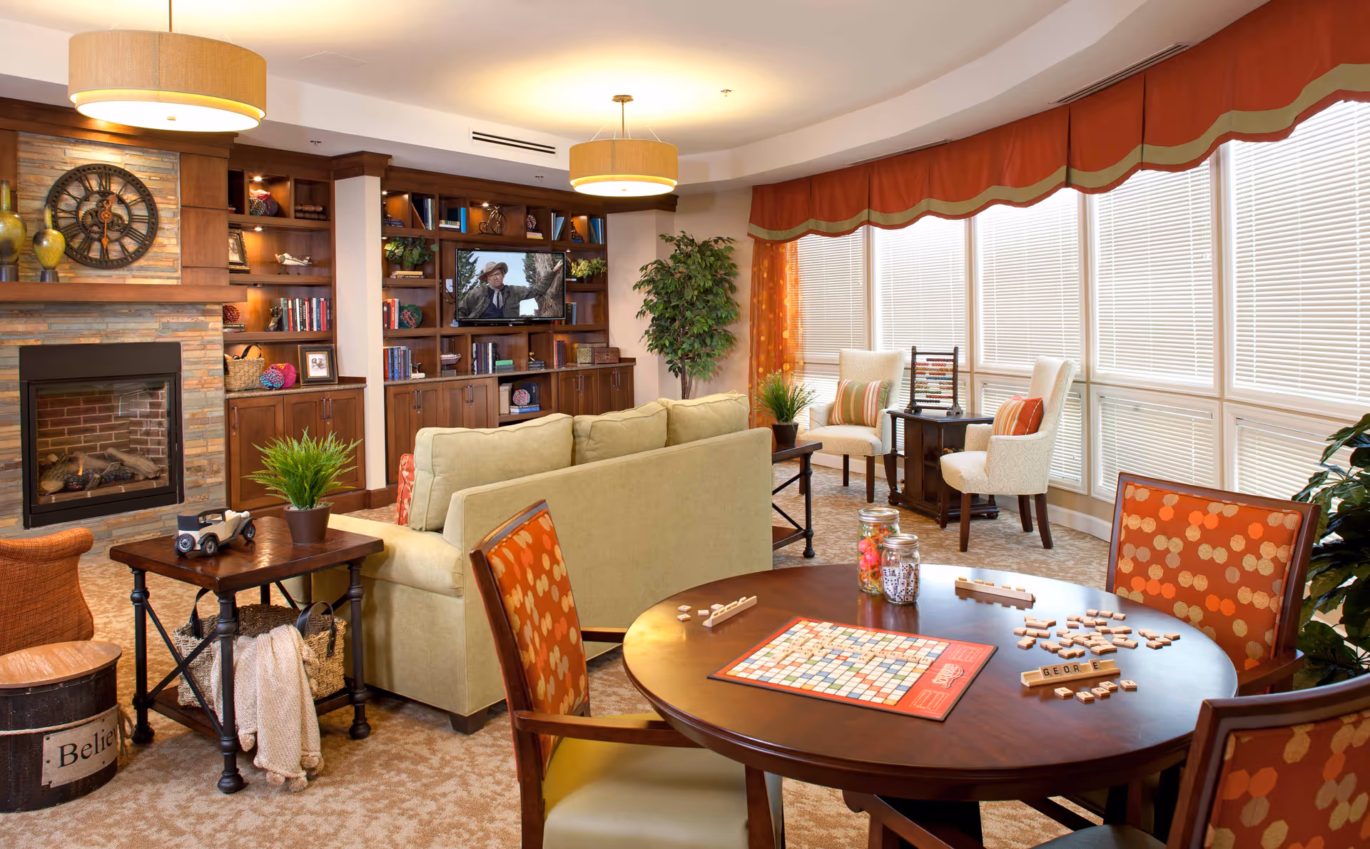 A cozy living room area with a beige sofa, wooden shelves filled with books and decorative items, a fireplace with a large clock above it, and a round table with a Scrabble game set up. The room is well-lit with large windows covered by blinds and orange valances, and there are several chairs with orange patterned upholstery around the table. Two beige armchairs with striped pillows are placed near the windows, and there are green plants adding a touch of nature to the space.