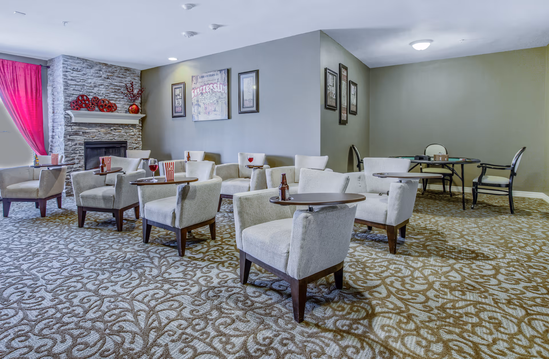 A lounge area with several beige armchairs each paired with small round tables. Some tables have drinks and popcorn containers on them. The room has a patterned carpet, a stone fireplace with decorative items on the mantel, framed artwork on the walls, and a bright pink curtain covering a window.