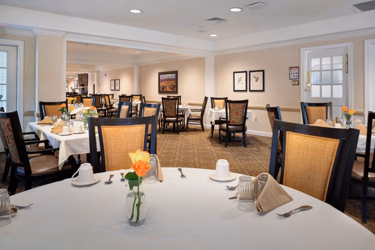 A dining room in a senior living facility with multiple tables covered in white tablecloths, set with cups, glasses, utensils, and beige napkins. Each table has a small vase with orange and yellow roses. The room has beige walls adorned with framed artwork and patterned carpet flooring. Wooden chairs with woven backs surround the tables.