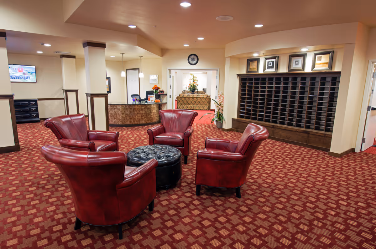 A cozy seating area in a senior living facility with four red leather armchairs arranged around a black tufted ottoman on a red patterned carpet. In the background, there is a reception desk with a computer and flowers, a wall clock, and a large wooden mail organizer with multiple slots. The space is well-lit with recessed ceiling lights and has beige walls.