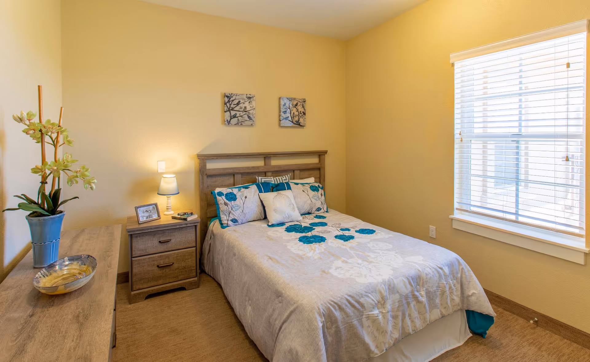 A cozy bedroom with a double bed featuring a floral bedspread and multiple pillows. There is a wooden nightstand with a lamp, a framed photo, and some books next to the bed. A wooden dresser with a decorative bowl and a potted plant is on the left side. The room has beige walls and a window with white blinds letting in natural light.