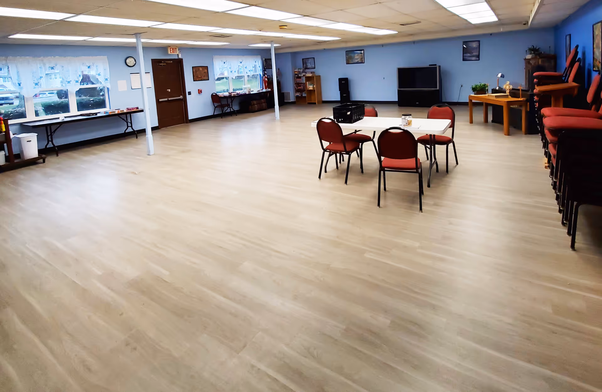 A large multipurpose room with light wood flooring and light blue walls. There are several red chairs stacked on the right side and a few chairs around a white table in the center. The room has windows along one wall, a door, a clock, and various pieces of furniture including tables and shelves. The ceiling has fluorescent lighting panels.