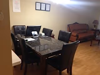 Interior view of a room with a dark marble dining table surrounded by six black chairs. In the background, there is a brown upholstered sofa and a small wooden side table with a lamp. The walls are light-colored, and there are framed documents or pictures hanging on the wall.