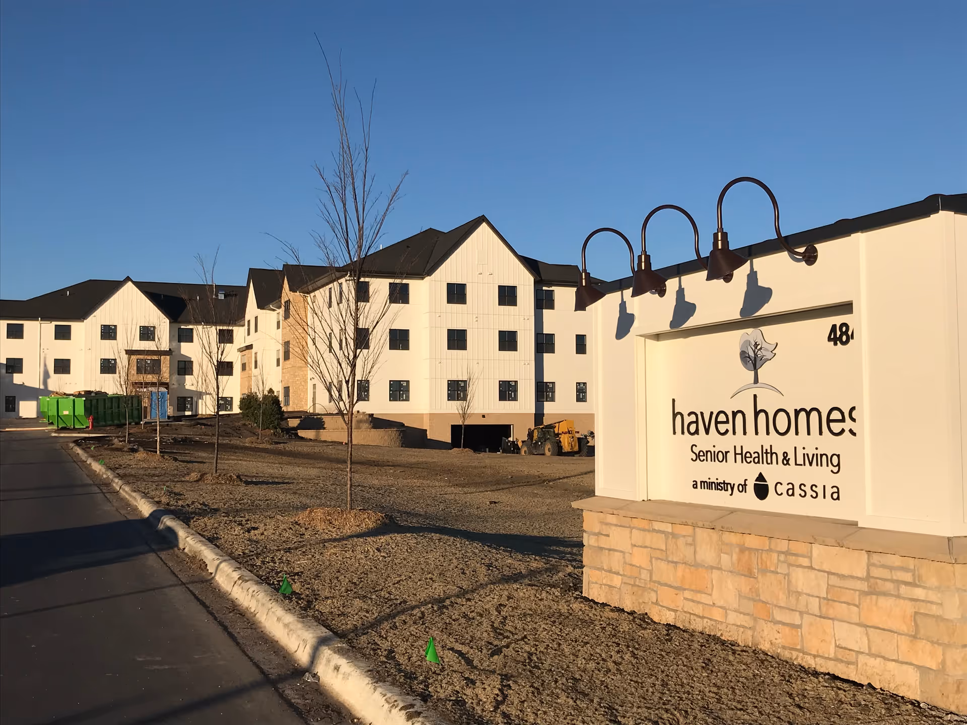 Exterior view of a senior living facility named Haven Homes with a large white building in the background and a stone sign in the foreground displaying the facility name and logo. The sky is clear and blue, and there are young trees planted along the sidewalk.