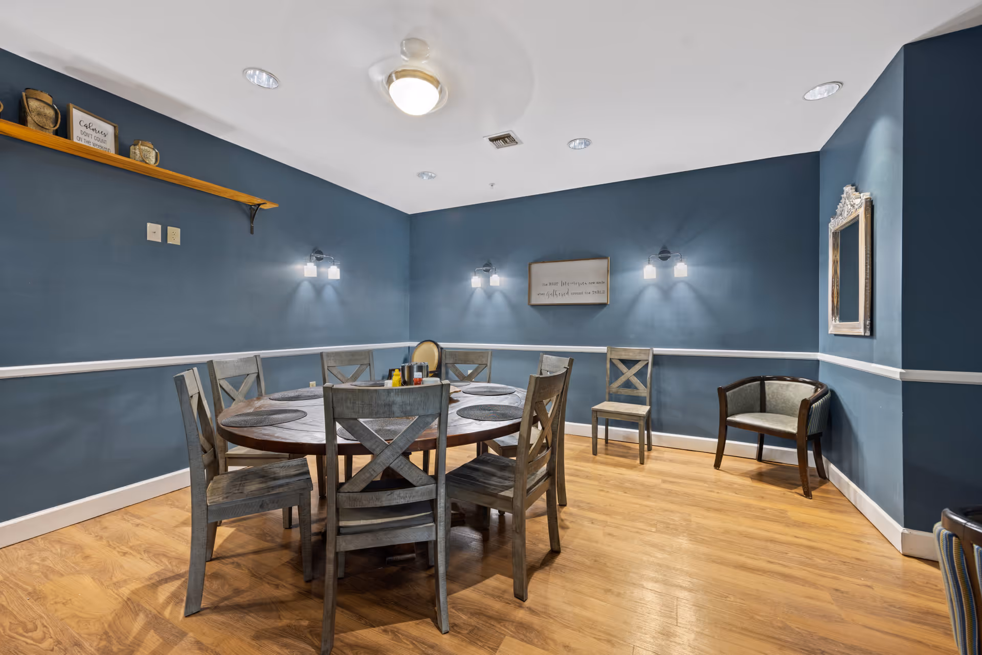 A dining room with a round wooden table surrounded by six wooden chairs with cross-back design. The walls are painted blue with white trim, and there are three pairs of wall-mounted lights. A small shelf with decorative items is mounted on the left wall, and a framed mirror is on the right wall. The floor is wooden, and there is a ceiling fan with a light fixture above the table.