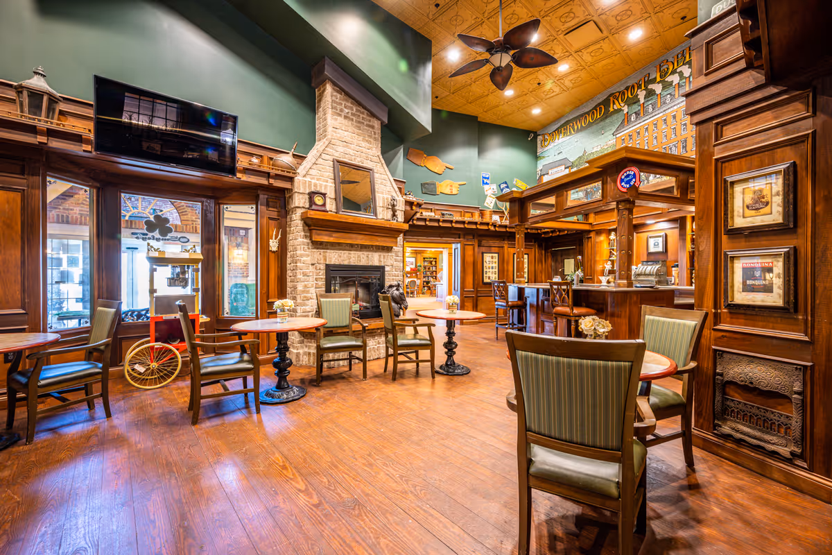 A cozy common area in Doverwood Skilled Nursing Facility featuring wooden floors, multiple round tables with chairs, a brick fireplace with a mirror above it, and a bar counter with stools. The walls are decorated with wood paneling, vintage signs, and a ceiling fan with leaf-shaped blades. Large windows allow natural light to enter the room.