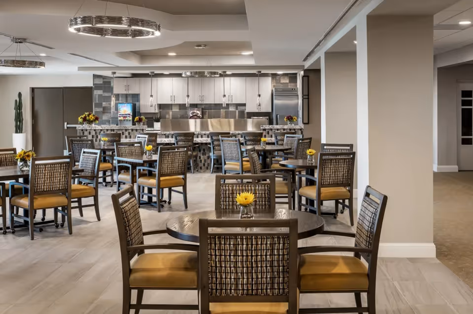 A modern dining area with multiple round tables and chairs featuring woven backs and cushioned seats. Each table has a small vase with a yellow flower. The background shows a kitchen area with white cabinets, stainless steel appliances, and a tiled backsplash. The room has a neutral color palette with beige walls and light flooring, and two circular ceiling light fixtures.