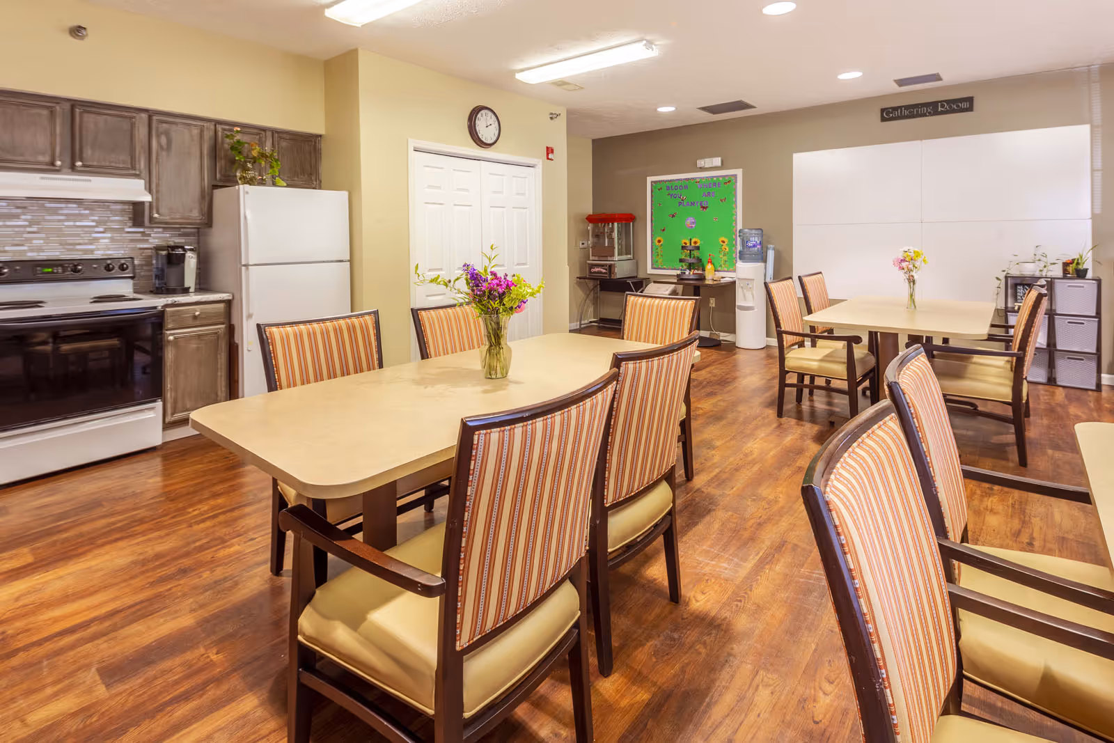 Communal dining area with multiple tables and striped chairs, a kitchenette with stove and refrigerator, and floral centerpieces.