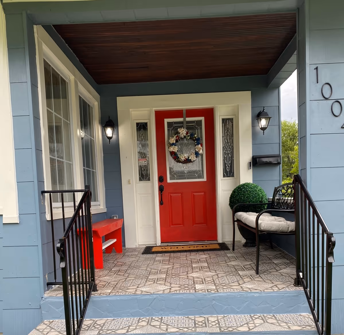 Front porch of a blue house with a red door decorated with a wreath, a small red bench and a cushioned chair.