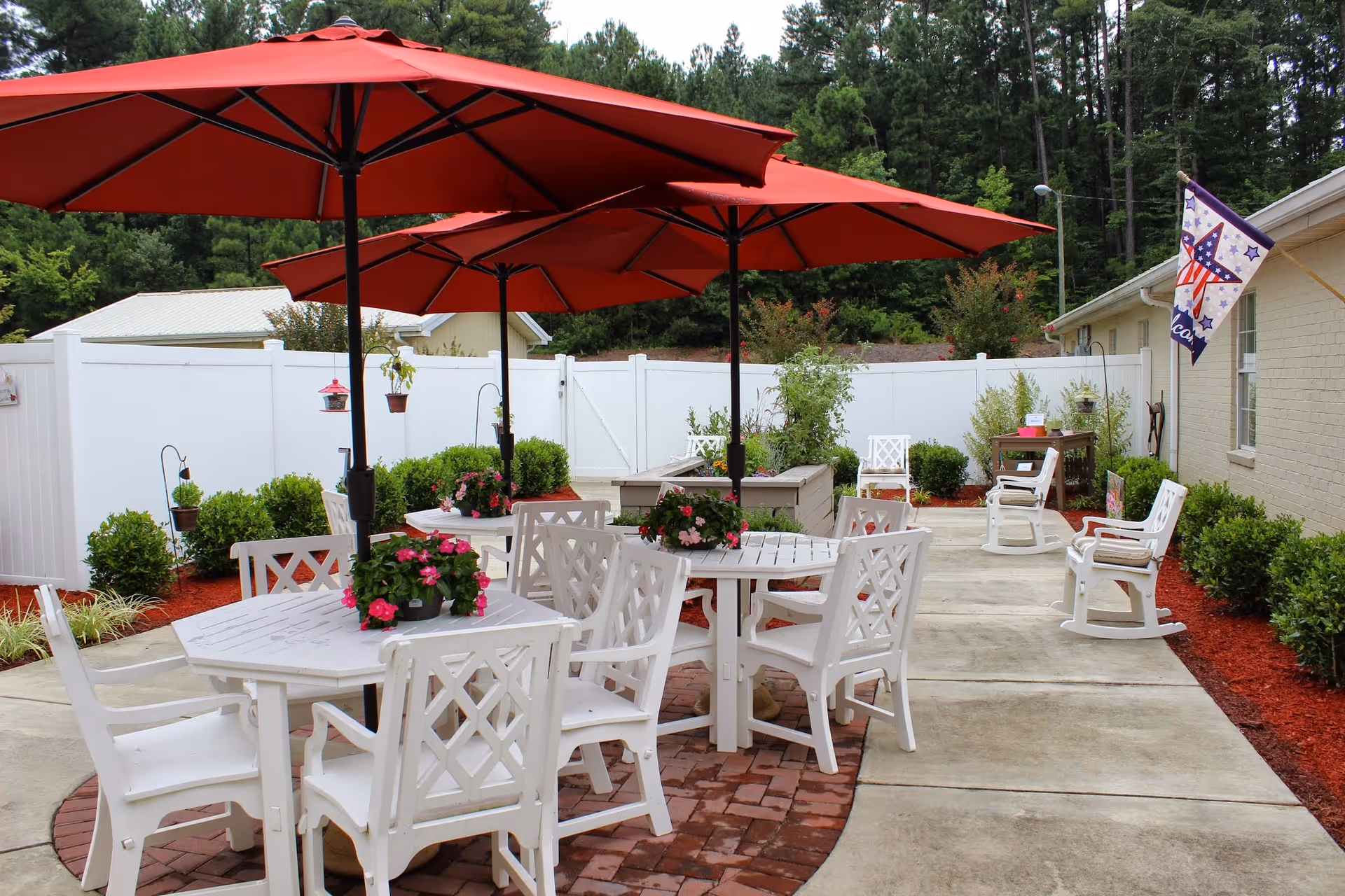 Patio seating area with white tables and chairs under red umbrellas, potted flowers, and a white fence.