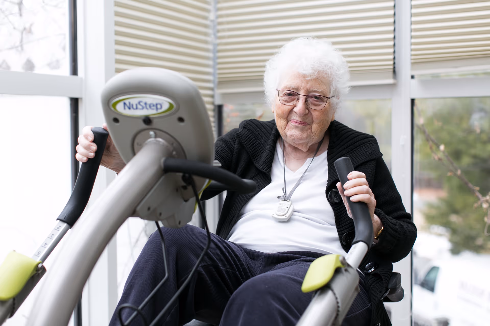 An elderly woman with white curly hair and glasses is exercising on a NuStep recumbent cross trainer indoors near large windows with blinds. She is wearing a black cardigan, white shirt, and dark pants, and has a medical alert device around her neck.
