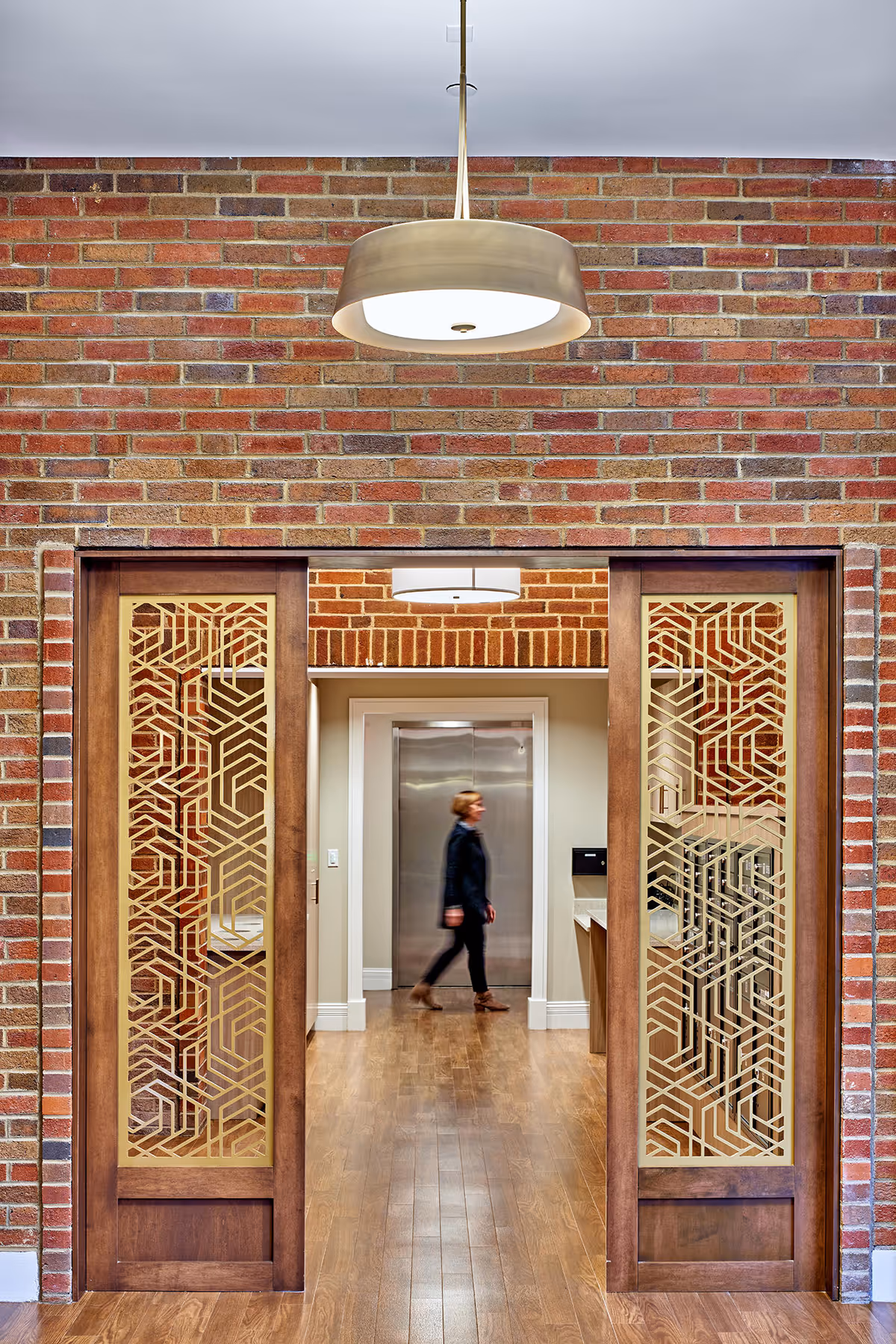 Interior hallway with decorative wooden lattice sliding doors in a brick wall, wood floors, a hanging light, and a person walking toward an elevator.