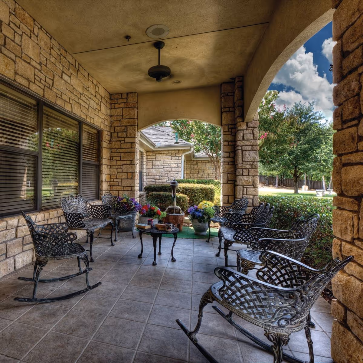 Covered outdoor patio area with stone walls and tiled floor, furnished with black metal rocking chairs and benches arranged around a small round table with flower pots and mugs. The patio overlooks a garden with bushes, trees, and a partly cloudy sky.