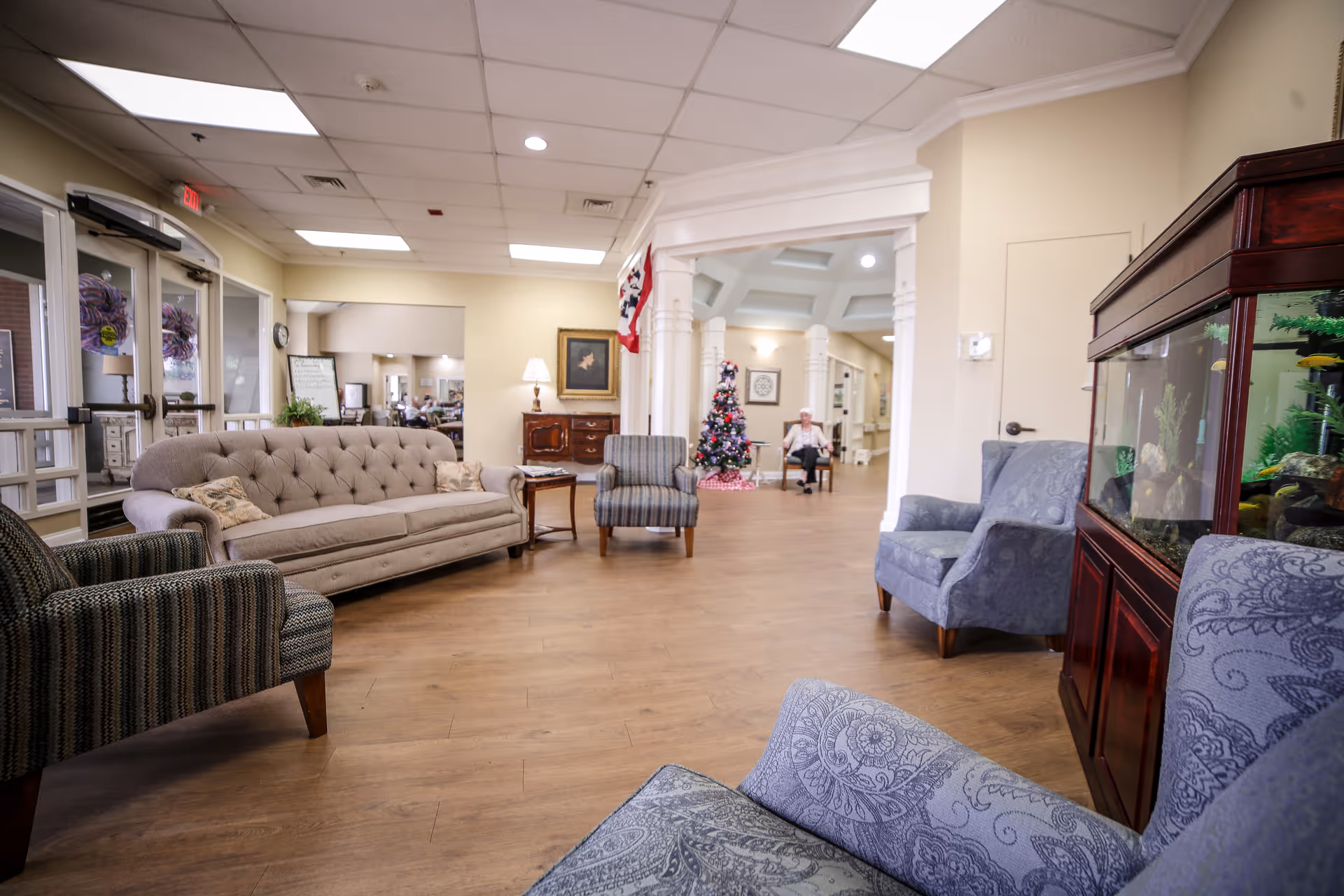 A spacious senior living facility common area with comfortable upholstered chairs and a sofa arranged around a wooden floor. There is a large fish tank on the right side and a decorated Christmas tree in the background near a seated elderly person. The room has a high ceiling with recessed lighting and large windows near the entrance on the left.