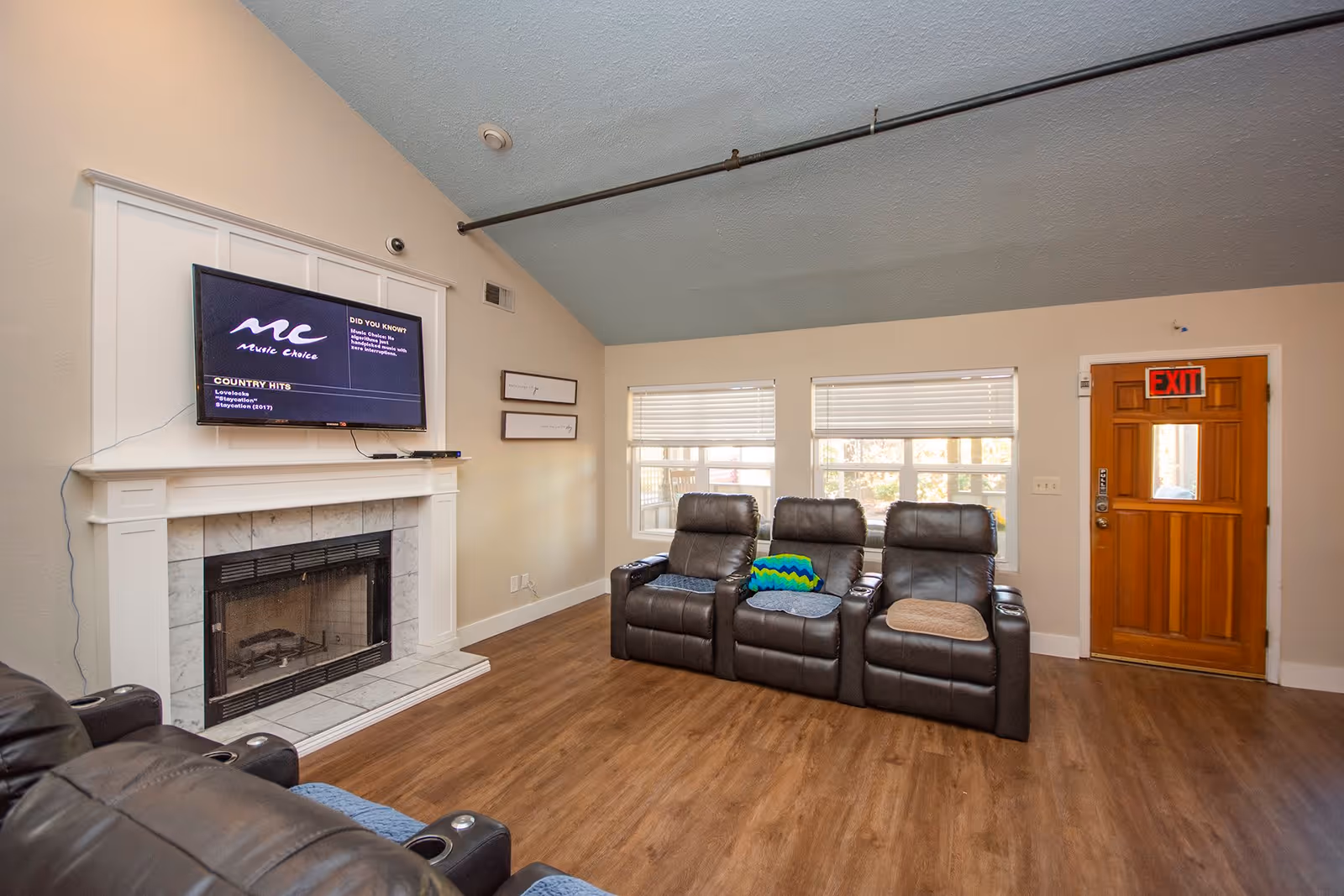 A common living area with three leather recliners facing a wall-mounted TV above a fireplace and a wooden exit door.