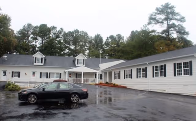 Exterior view of a single-story white building with black shutters and a dark roof, surrounded by trees. A black car is parked on the wet asphalt driveway in front of the building.