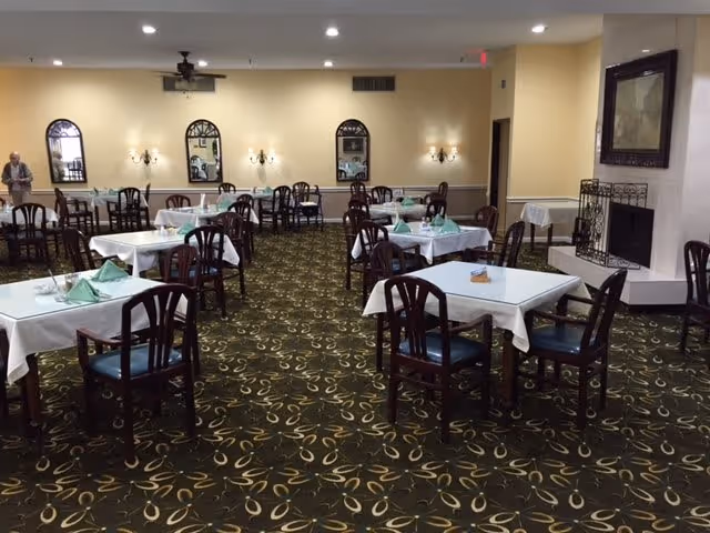 Dining room with multiple tables covered in white tablecloths and green folded napkins, wooden chairs, patterned carpet, and wall mirrors.