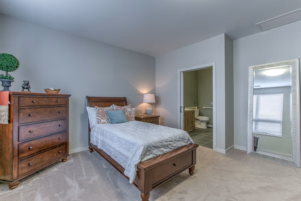 Well-lit bedroom with a wooden bed and dresser, a nightstand lamp, and an open doorway leading to an en-suite bathroom.